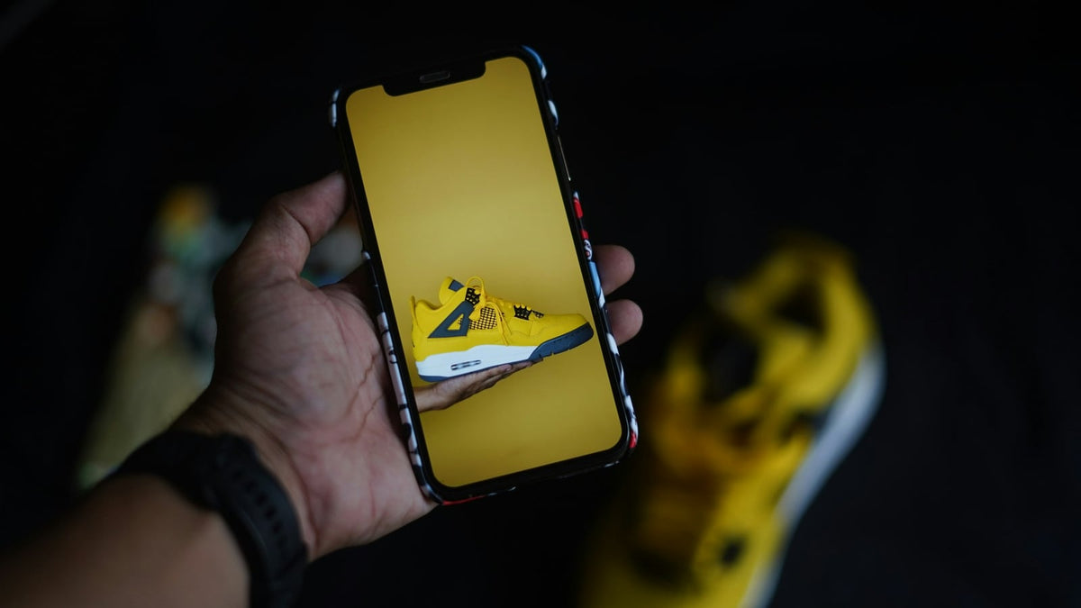 A hand holding a phone showing a sneaker listing, with a sneaker beside it