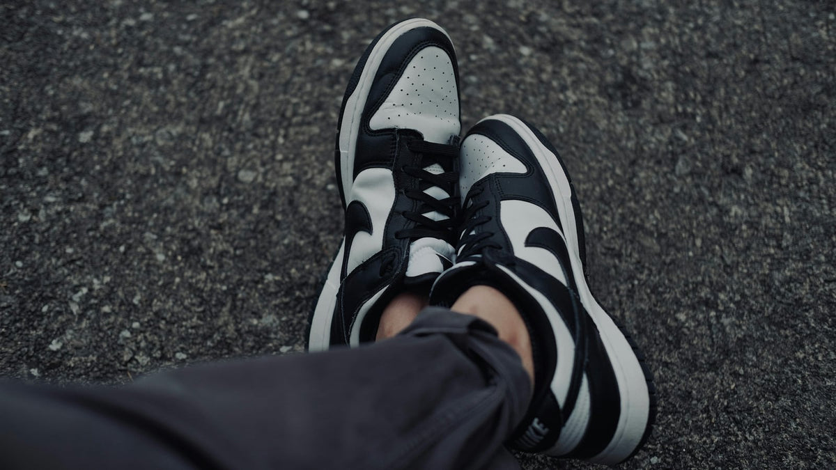 Someone wearing a black and white Nike Dunk Low Panda, photographed looking down at asphalt