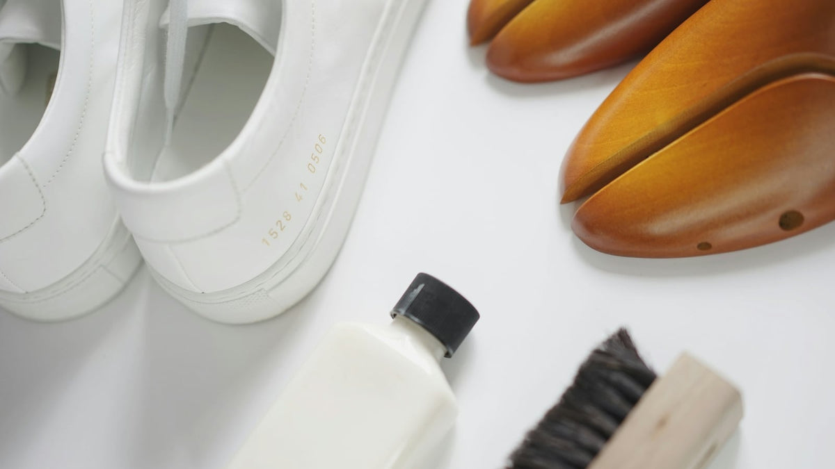 White leather sneakers laid out beside wooden shoe trees, a bottle of cleaner and a suede brush